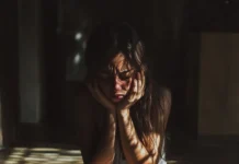 Trauma Religioso: Supera las Secuelas y Recupera tu Vida Una mujer joven sentada en el suelo, con las manos sosteniendo su rostro en un gesto de angustia y tristeza, iluminada por rayos de luz que atraviesan una ventana. Representa las secuelas emocionales del trauma religioso.